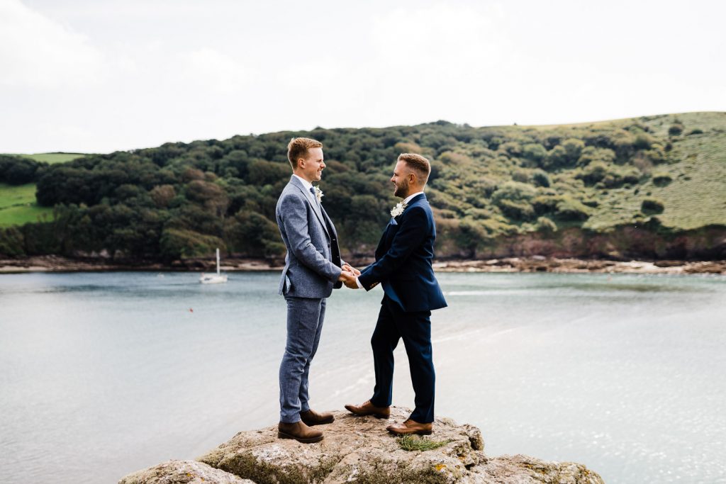 Grooms hold hands on rock next to the River Yealm. 