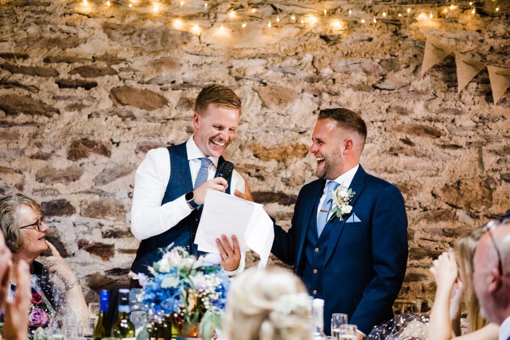 Grooms laugh during speeches in stone barn.