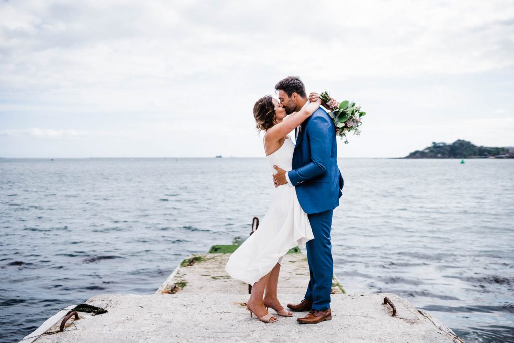Bride and groom kiss on Plymouth waterfront, in front of Plymouth Sound and Drakes Island.