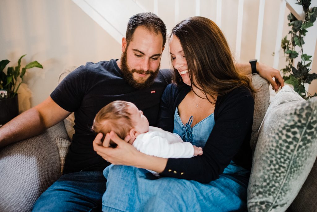 new parents smile at their baby daughter whilst seated on sofa.