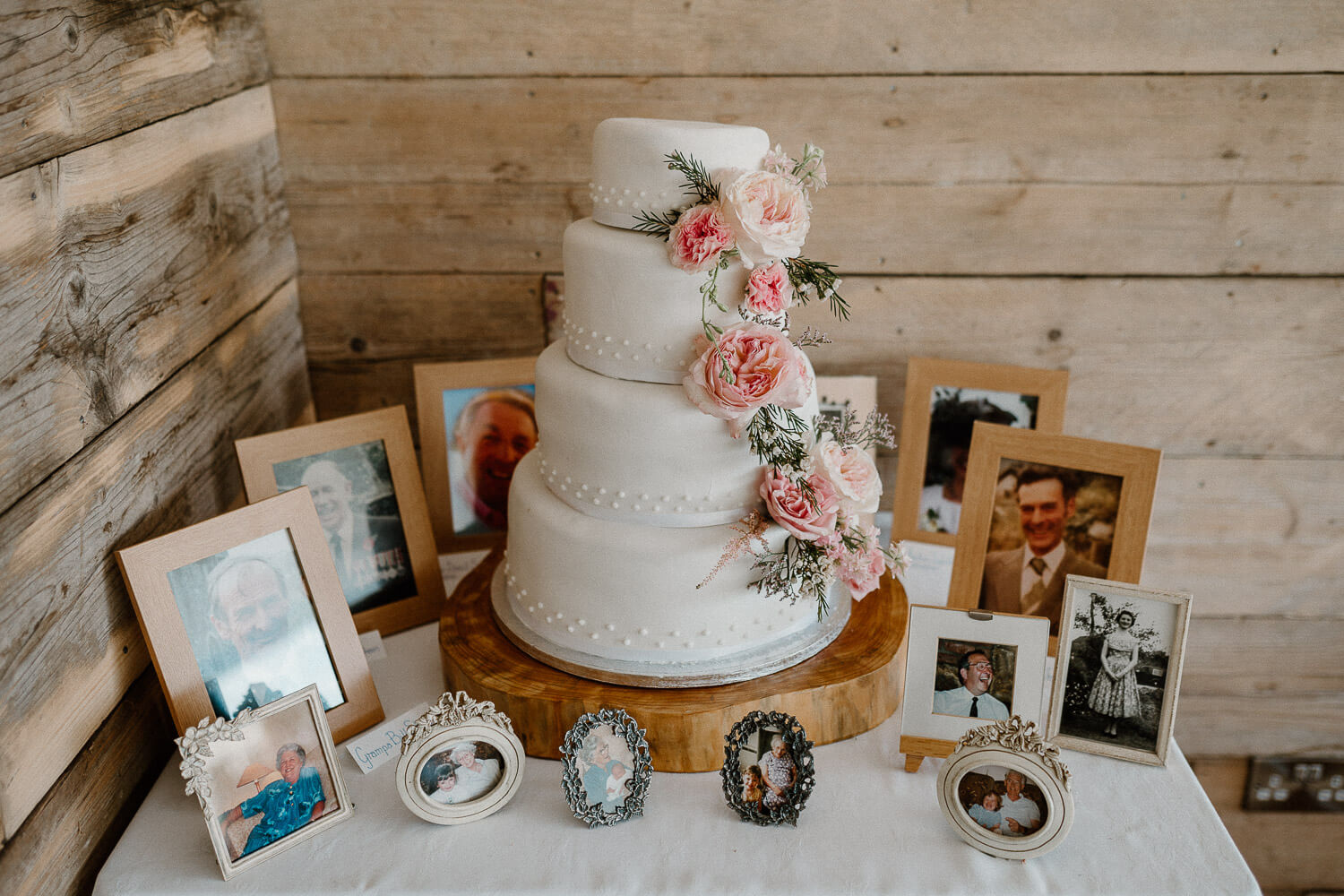 White tiered wedding cake decorated with pink flowers and surrounded with photos of loved ones departed.