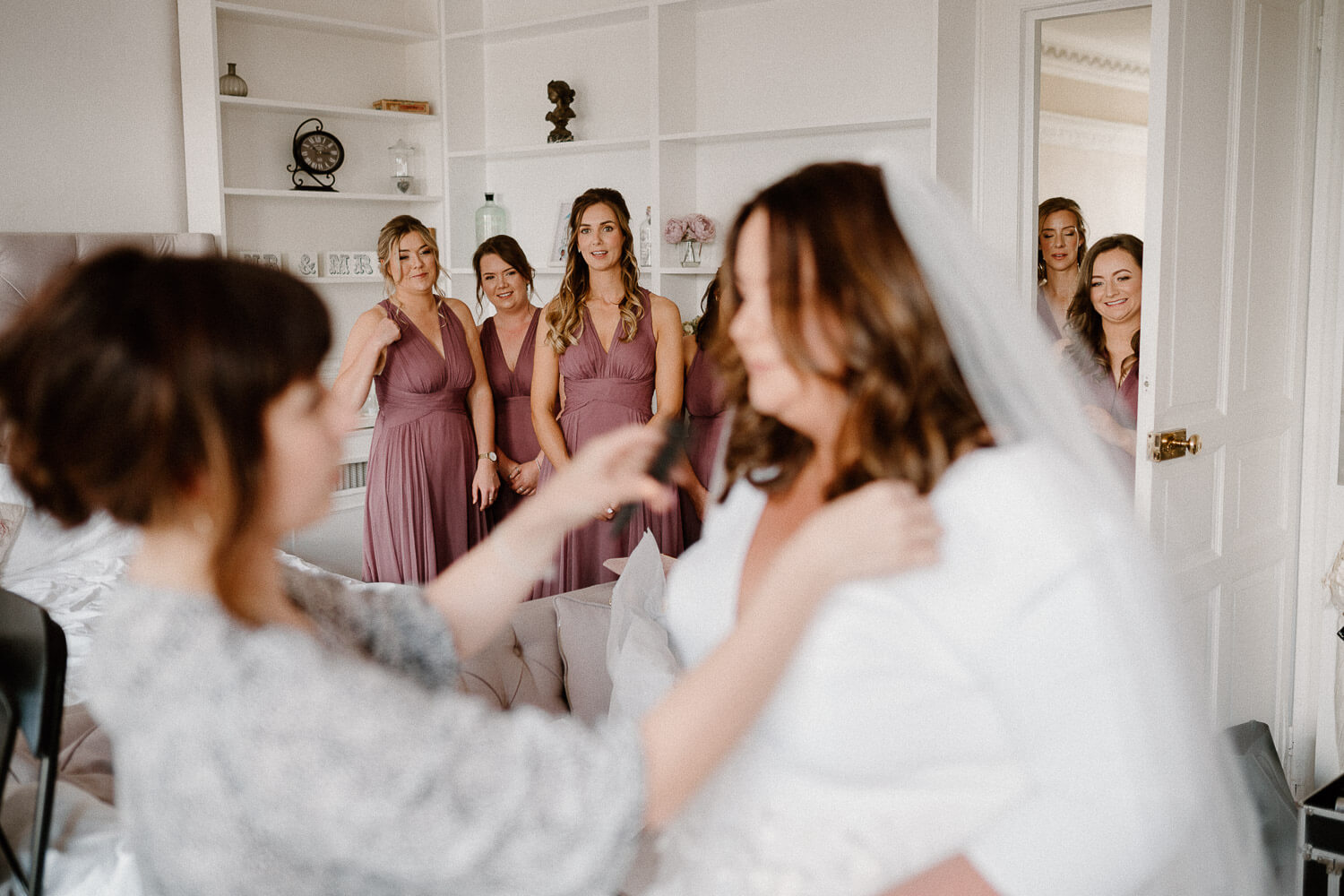 Hair stylist makes finishing touches to bride as bridesmaids look on.