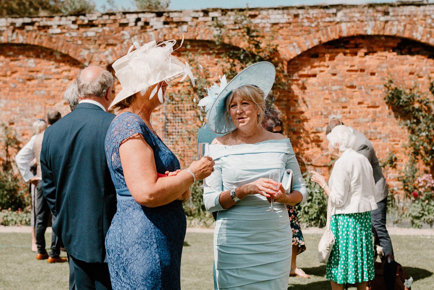 Female wedding guests wearing hats enjoy reception drinks in the walled gardens at Kingston Estate, Devon.