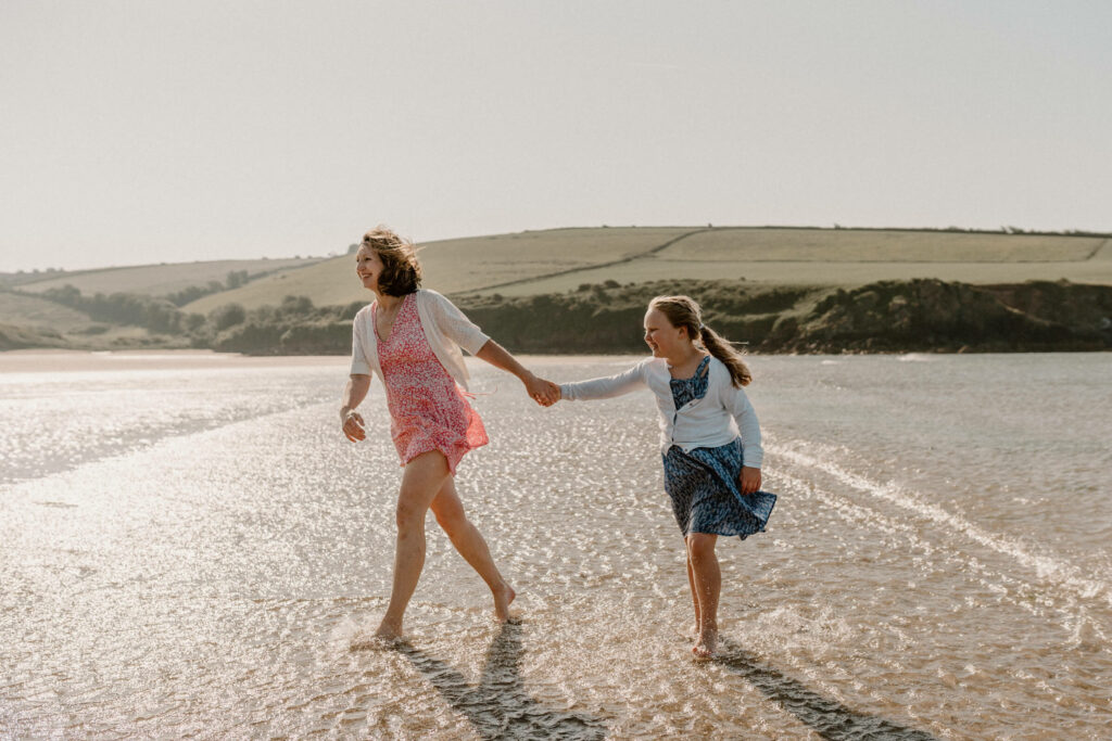 mother and daughter walk through shallow water on beach, hand in hand