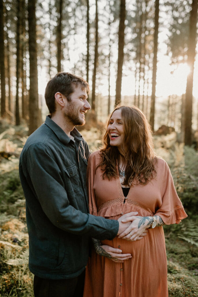 Expectant parents hold hands over their bump in the woods.
